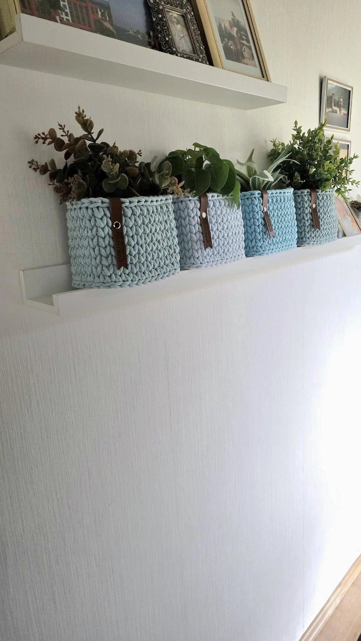 Gehäkelte Mini-Körbchen auf einer schmalen Regalfläche, mit braunen Lederlabels als Kontrast zur blauen Baumwollschnur.