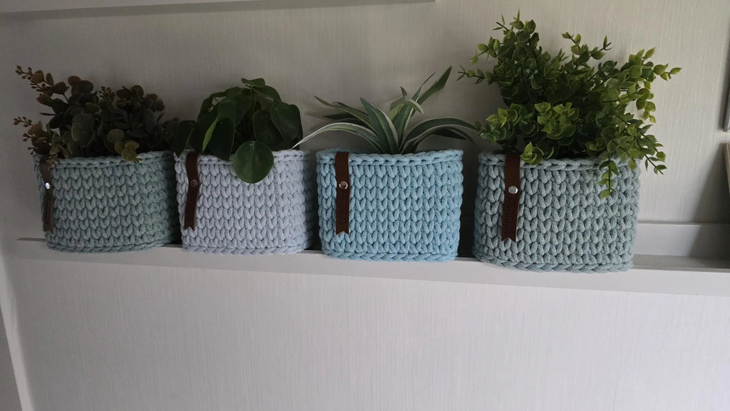 Hellblaue und graublaue Mini-Körbchen mit Lederlabels, deren obere Ränder von dekorativem Pflanzengrün umgeben sind.