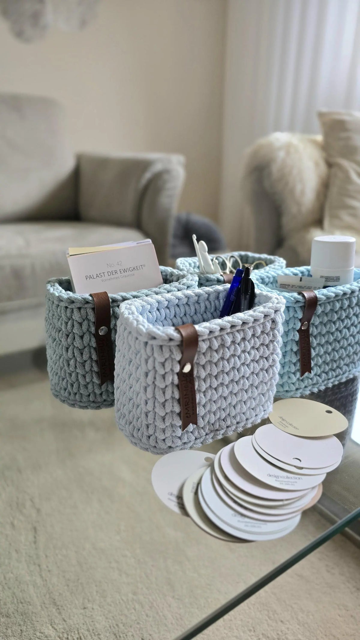 Gehäkelte Mini-Körbchen auf einer schmalen Regalfläche, mit braunen Lederlabels als Kontrast zur blauen Baumwollschnur.